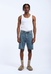 Shorts di jeans di un blu chiaro, di lunghezza media, con una vestibilità standard. Abbinati a una canottiera bianca e a sandali beige, completati da calze lavorate a maglia.