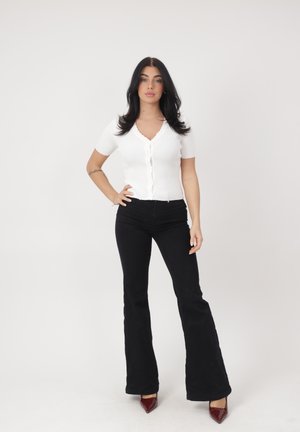 Vrouw met lang donker haar staat op een witte achtergrond, gekleed in een wit kort mouw shirt met knopen, zwarte wijde broek en bordeauxrode puntige hakken.