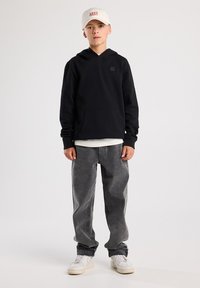 Felpa nera con cappuccio e tasca frontale, jeans grigi e scarpe da ginnastica bianche. Il cappellino è di colore chiaro con testo rosso. Design semplice.