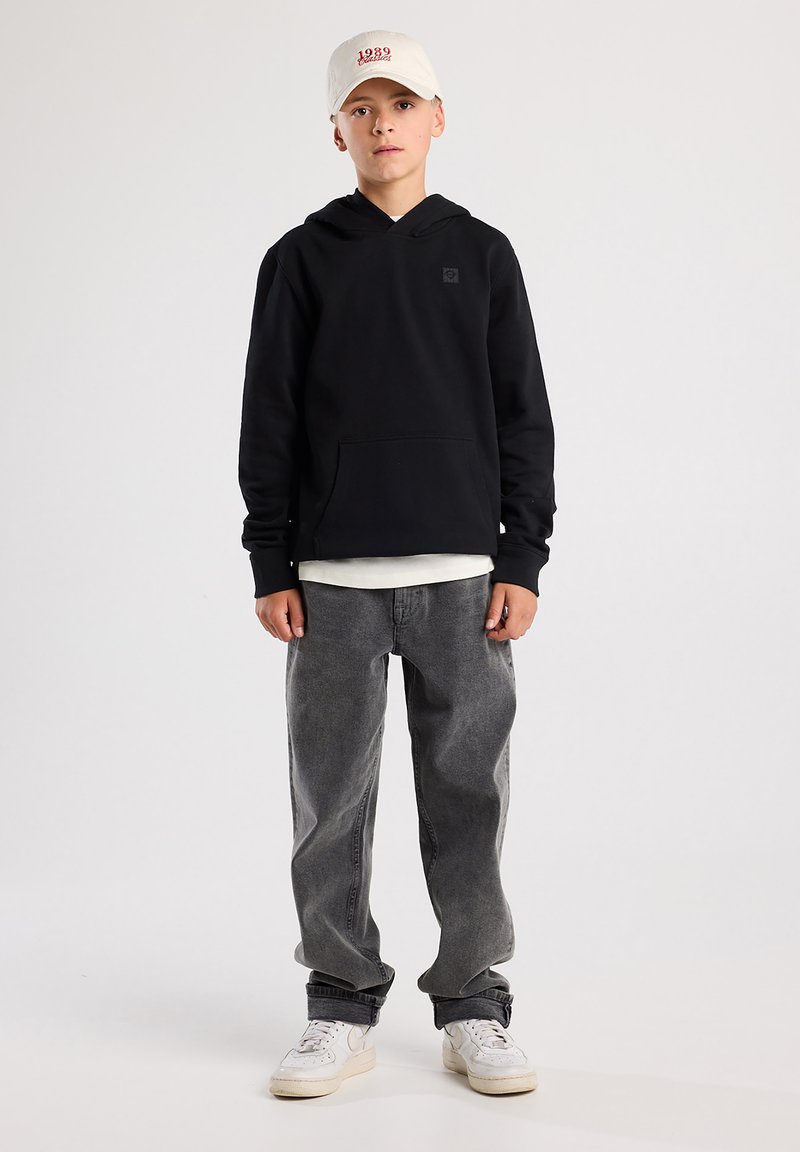 Felpa nera con cappuccio e tasca frontale, jeans grigi e scarpe da ginnastica bianche. Il cappellino è di colore chiaro con testo rosso. Design semplice.