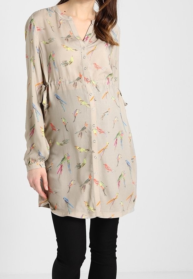 Tunique beige à manches longues avec des imprimés d'oiseaux colorés, boutonnée devant, portée sur un pantalon noir moulant par une femme debout.