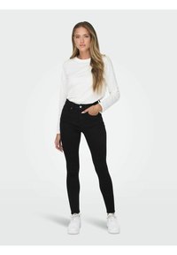 Ženska stoji oblečena v belo srajco z dolgimi rokavi, črne skinny kavbojke visoke pasu in bele superge, z eno roko v žepu.