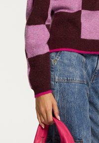 Karirana jopica iz roza in maroon volne, z ribanim ovratnikom in manšetami. Roka drži roza usnjeno torbico. Vidni so jeans v denim materialu.