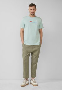 Jasno-miętowa bawełniana koszulka z okrągłym dekoltem i grafiką "JAWS" w kolorze czerwonym i niebieskim. Połączona z oliwkowymi spodniami w tapered i beżowymi sneakerami.