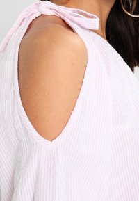 Vaaleanpunainen, teksturoitu kangastoppi, jossa on kylmät olkapäät, sitomisnauha ja hienovaraiset valkoiset raidat.