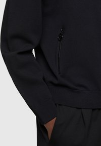 Felpa a maglia scura con una vestibilità comoda, polsini a coste e una tasca con zip sul lato. Il materiale ha una texture liscia.