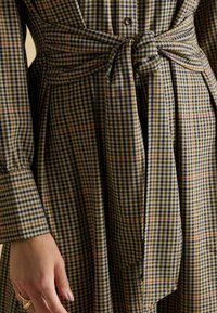 Brūna un zila rūtainā kleita ar sasietu jostas detaļu un pogu aizdari, ar garām piedurknēm un mīkstu auduma tekstūru.