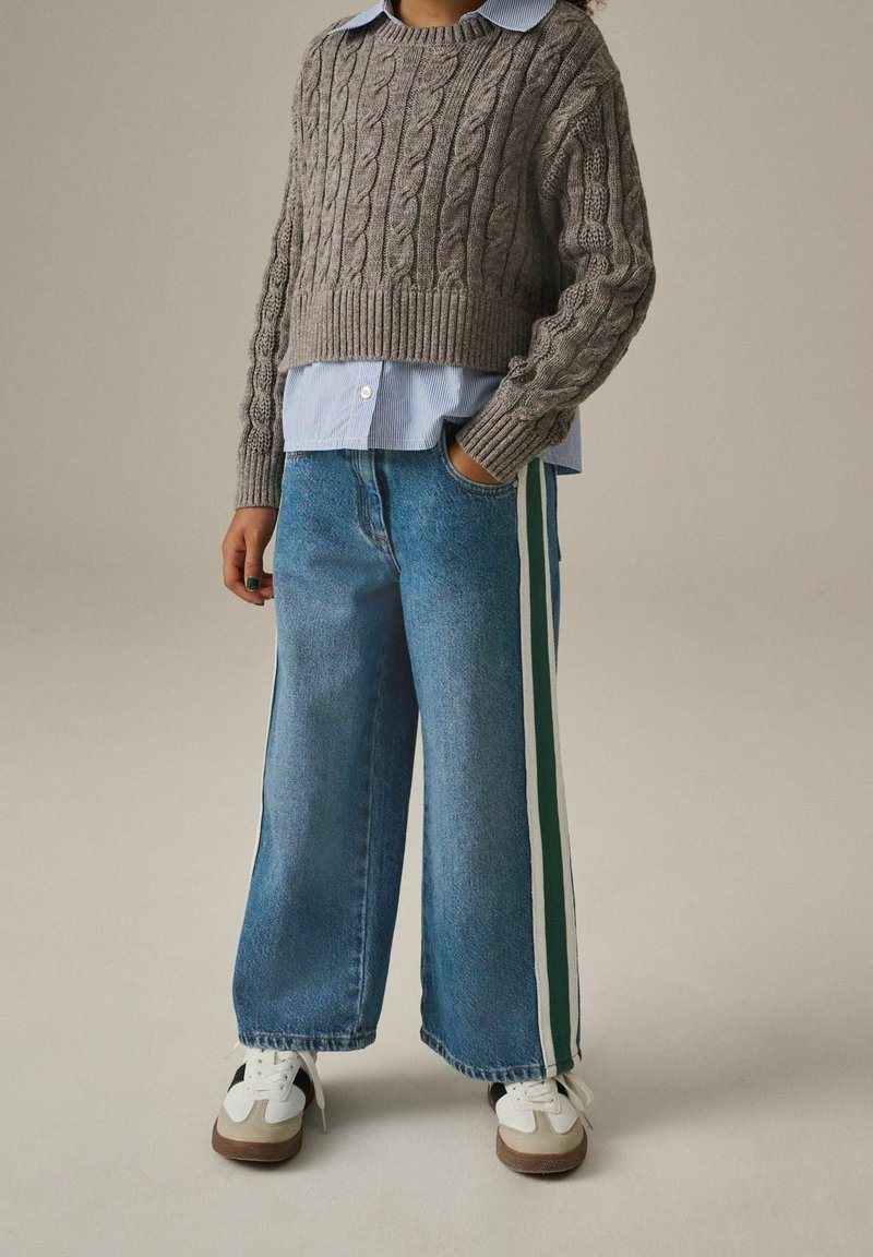 Personne portant un pull gris à torsades par-dessus une chemise bleue à boutons, un jean à jambes larges avec des bandes latérales vertes et blanches, et des baskets blanches, debout avec la main dans la poche.