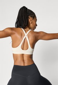 Bēša sporta krūšturis ar krustenisku aizmugures dizainu, izgatavots no gludas auduma. Kombinēts ar melnām, augsta vidukļa legingiem.