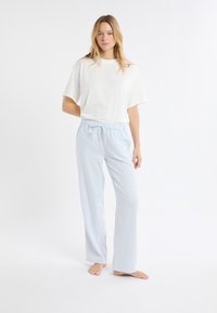 Biała bawełniana koszulka zestawiona z jasnoniebieskimi, w kratę, piżamowymi spodniami z elastycznym paskiem i sznurkiem, noszona boso na białym tle.
