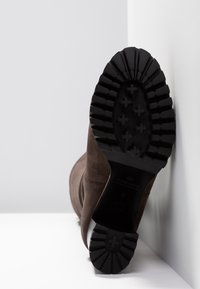 Botte en daim marron reposant sur le côté, montrant la semelle noire robuste avec des rainures profondes et des motifs en forme de croix.