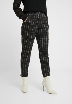 Vrouw die zwart-witte geruite broek draagt met witte enkellaarsjes met hak en een zwarte top met lange mouwen, handen in de zakken.