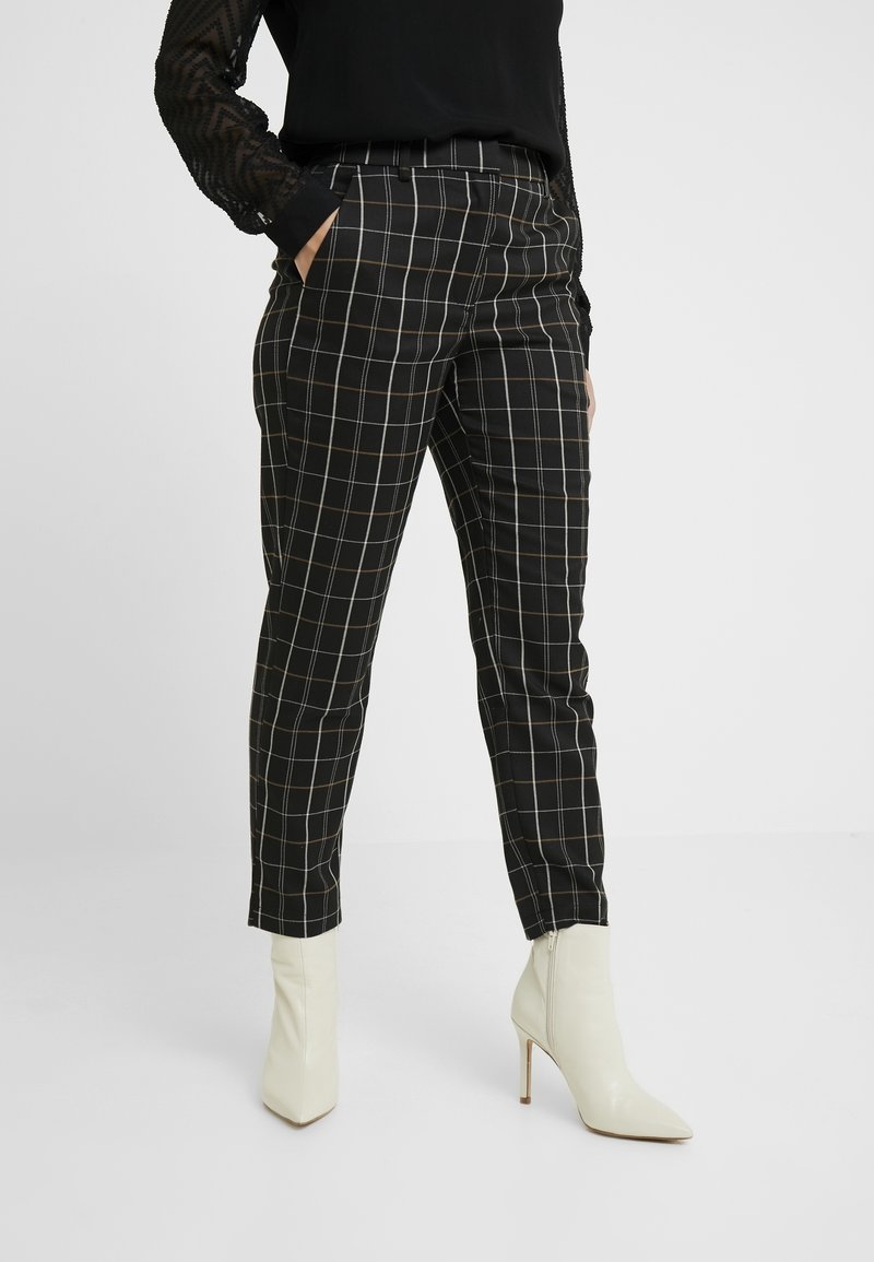 Femme portant un pantalon à carreaux noir et blanc avec des bottines blanches à talons et un top noir à manches longues, mains dans les poches.