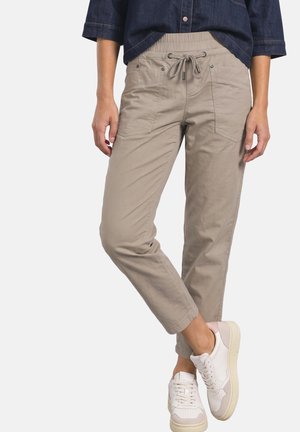 Person iført beige bukser med snor, hvide sneakers og en mørkeblå skjorte med hænder afslappet langs siderne.