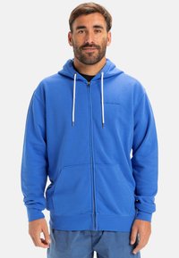 Blue zip-up hoodie made of soft, textured fabric. Features a drawstring hood, front pockets, and logo detail on the chest.