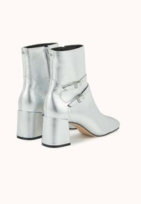 Bottes chevilles métalliques argentées avec un bout pointu et un talon carré, dotées de deux boucles ajustables et d'une fermeture éclair latérale. Texture lisse et brillante.