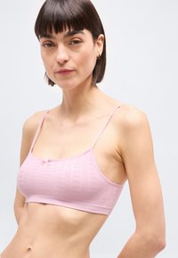 Γυναίκα με κοντά σκουρόχρωμα μαλλιά, ντυμένη με ανοιχτό ροζ bralette με δαντελωτό μοτίβο και λεπτές τιράντες, σε ουδέτερο ανοιχτό φόντο.