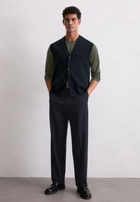 Dunkelgrüne Strickweste über einem olivgrünen Langarmshirt, kombiniert mit schwarzen maßgeschneiderten Hosen und schwarzen Schuhen. Einfache Knöpfe an der Weste.