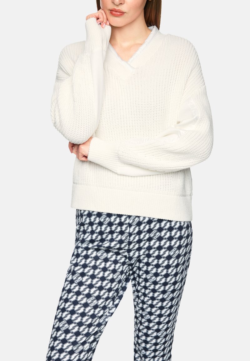 Weißer gestrickter Pullover mit V-Ausschnitt, langen Ärmeln und strukturiertem Muster. Kombiniert mit marineblauen, gemusterten Hosen mit ineinandergreifenden Formen.