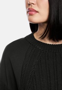 Schwarzer Pullover mit strukturiertem Strickdesign und geripptem Kragen; glattes Material am Körper; Nahaufnahme von Kragen und Schulter.