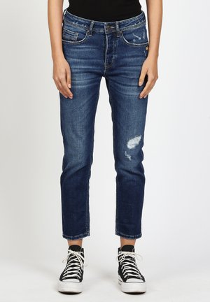 Person trägt abgenutzte dunkelblaue Jeans und schwarze High-Top-Sneaker, steht vor einem schlichten weißen Hintergrund.