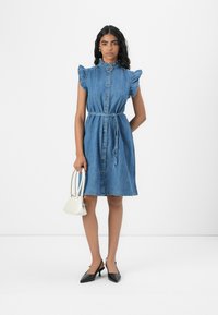 Denim ruha rövid fodros ujjakkal, gombos elülső résszel, megkötött derékkal, térd alá ér. Fehér kézitáskával és fekete magassarkúval viselve.