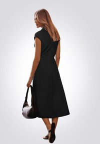 Vestido negro de manga corta con un cuerpo ajustado y falda acampanada, de tela texturizada. La persona lleva un bolso oscuro de hombro y sandalias planas.