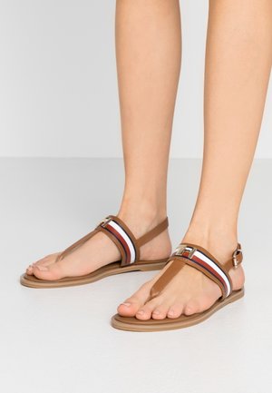 Stopy ubrane w brązowe sandały na płaskiej podeszwie z białymi, czerwonymi i niebieskimi paskami oraz złotym zapięciem na gładkiej białej podłodze.