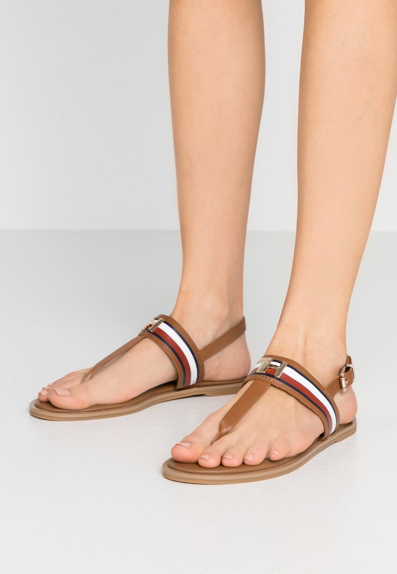 Stopy ubrane w brązowe sandały na płaskiej podeszwie z białymi, czerwonymi i niebieskimi paskami oraz złotym zapięciem na gładkiej białej podłodze.