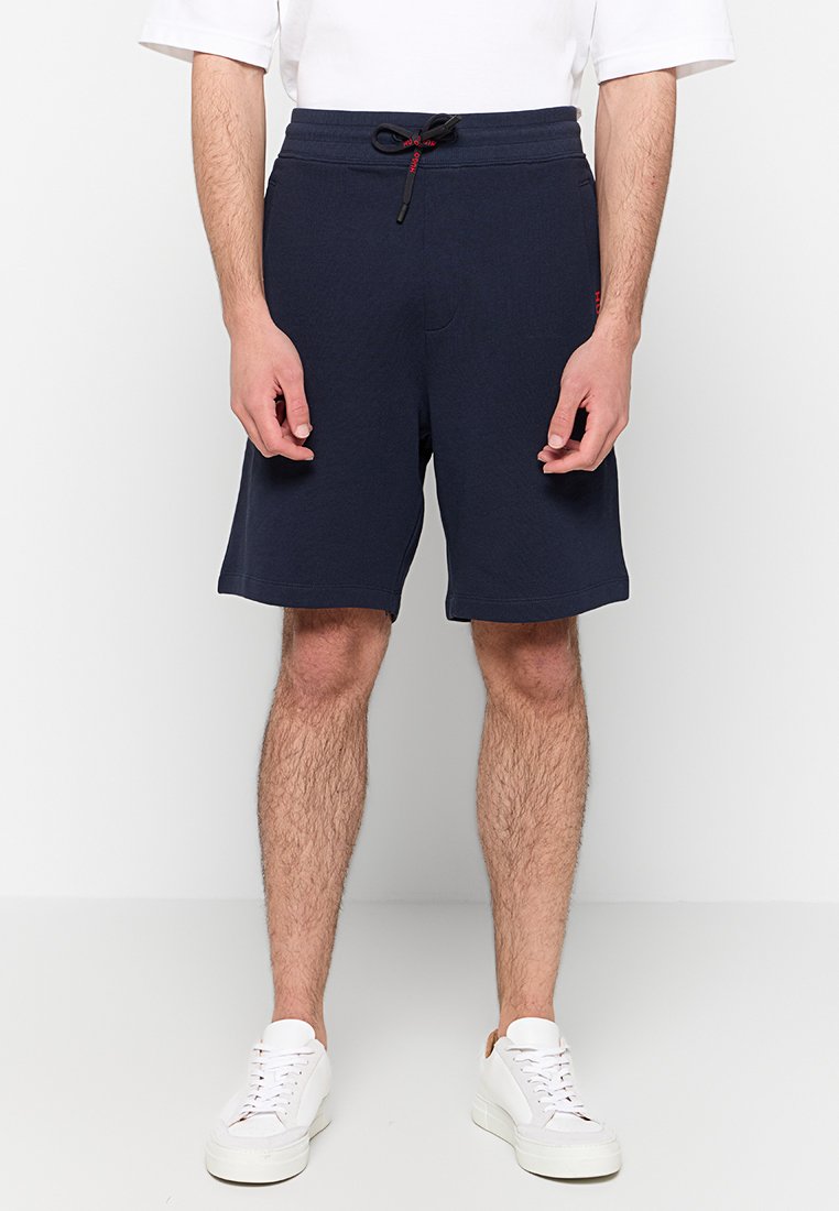 Dunkelblaue Shorts mit elastischem Bund und Kordelzug, ausgestattet mit Seitentaschen und einer glatten Baumwolltextur. Getragen mit weißen Turnschuhen.
