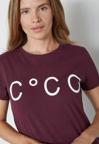 Moteris su šviesiai rudais plaukais, vilkinti tamsiai raudoną marškinėlį su dideliais baltais rašmenimis "C ° C O" priekyje, stovi prieš paprastą sieną.