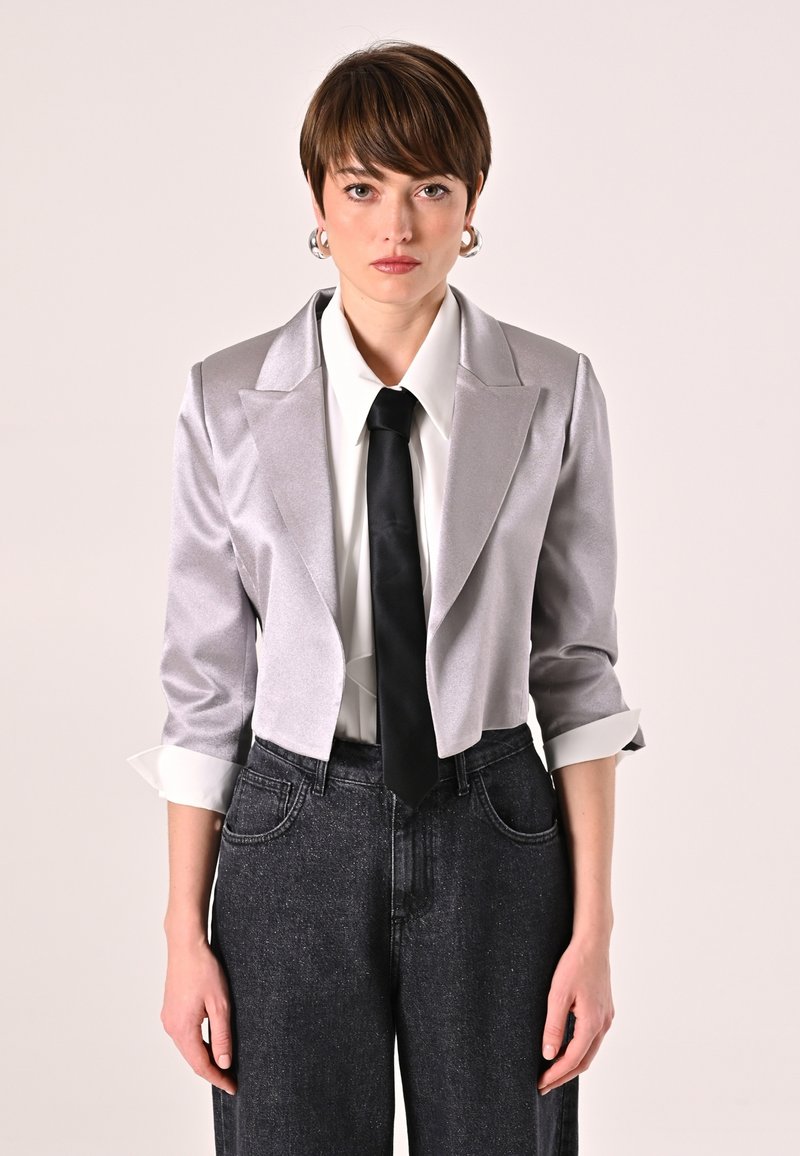 Blazer corto in satin argento con rever a punta, abbinato a una camicia bianca e una cravatta nera. Indossato con pantaloni in denim scuro a gamba larga.