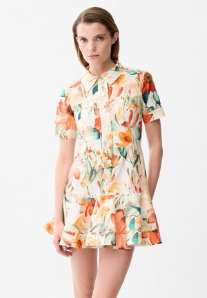 Donna che indossa un vestito floreale a maniche corte con colletto, fiori arancioni, verdi e gialli, con bottoni sul davanti e gonna svasata.