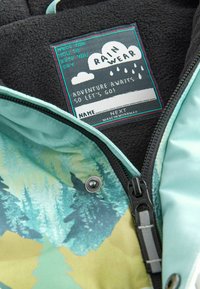 Étiquette sur une veste en polaire noire avec fermeture éclair bleue, texte "VÊTEMENT DE PLUIE" sous un graphique de nuage de pluie, et tissu imprimé forêt partiellement visible en dessous.