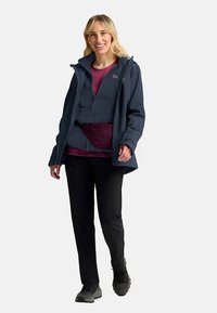 Navy waterproof jacket with a hood, layered over a burgundy zip-up fleece and dark pants; model wearing grey hiking shoes.