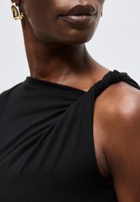 Κοντινό πλάνο ενός ατόμου που φοράει μαύρο αμάνικο τοπ με λεπτομέρεια δεμένη στον ώμο και ένα χρυσό σκουλαρίκι σε αφηρημένο σχήμα.