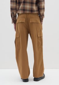 Braune Cargo-Hosen mit lockerer Passform, ausgestattet mit zwei seitlichen Taschen und Gesäßtaschen. Kombiniert mit einem karierten Hemd. Glatte Baumwolltextur.