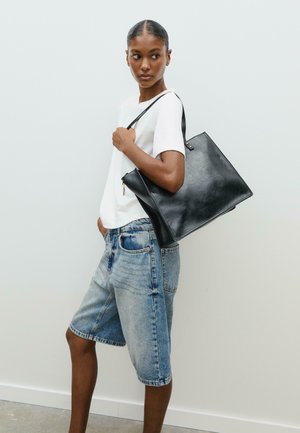 Sac en cuir noir à porter à l'épaule avec un design structuré, doté d'une fermeture éclair et d'une bandoulière, placé sur un fond neutre.