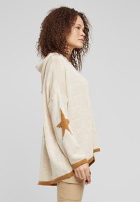 Light beige knitted hoodie with a large brown star design on the left sleeve, featuring a relaxed fit and ribbed hem.