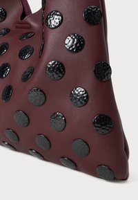 Bolsa de mão em couro bordô com rebites circulares pretos em relevo. Apresenta uma superfície macia e texturizada, com uma forma triangular que realça o interesse visual.