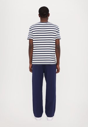 Homme debout, dos à nous, portant une chemise à manches courtes blanche et bleu marine à rayures, un pantalon bleu marine et des chaussures blanches sur un fond uni.