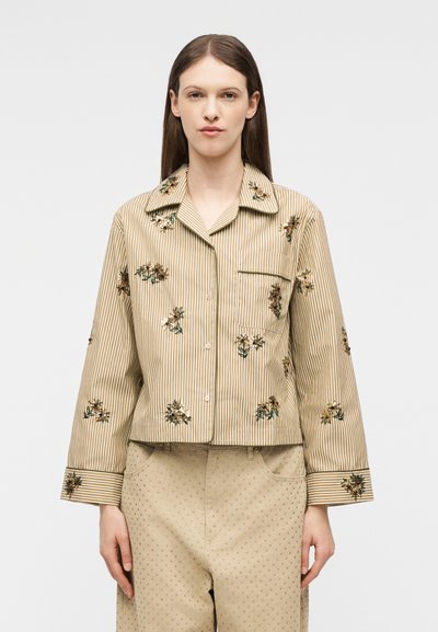 Γυναίκα με πουκάμισο μπεζ με ρίγες και κεντήματα με λουλούδια, και μπεζ παντελόνι με μικρά πουά, στέκεται μπροστά σε απλό φόντο.