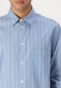 Camisa de botones a rayas azules con botones blancos, cuello y un bolsillo en el pecho, hecha de tejido de algodón ligero.