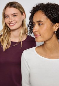 Twee vrouwen dragen langemouwen shirts: de een in donkerpaars, de ander in wit. Beide shirts hebben een zachte, gladde textuur en ronde halslijnen.