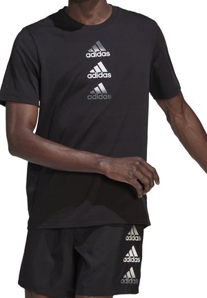 Hombre vestido con una camiseta negra de Adidas y pantalones cortos, cada uno con tres logotipos verticales plateados de Adidas en el centro frontal y en la pierna izquierda.