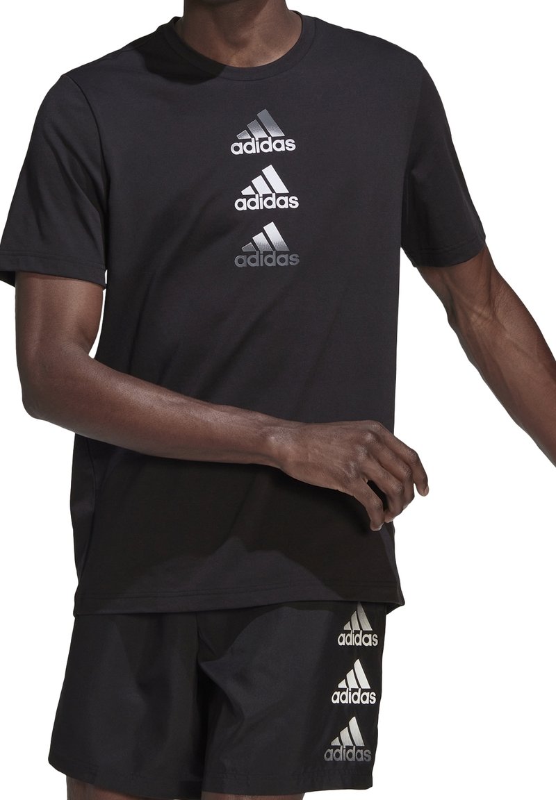 Muž v černém tričku a šortkách Adidas, na kterých jsou vpředu uprostřed a na levé noze tři vertikální stříbrné loga Adidas.