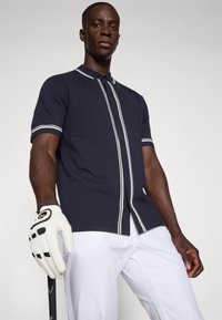 Polo azul-marinho com riscas brancas, mangas curtas e fecho de botão. Usado com calças de golfe brancas e segurando um taco de golfe com uma luva.