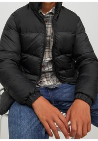 Osoba siedząca, ubrana w czarną puchową kurtkę na beżowej koszuli w kratę zapinanej na guziki i niebieskich dżinsach, z rękami spoczywającymi na kolanach.