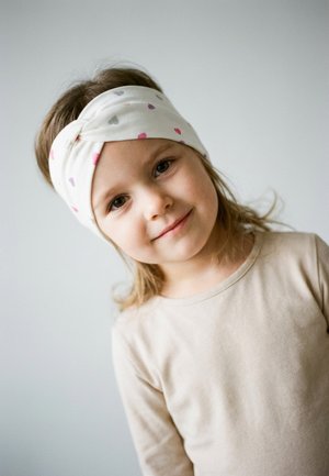 Young girl with light brown hair wearing a beige shirt and white headband with small colorful hearts, smiling slightly against a plain background.