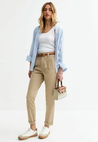 Beige maßgeschneiderte Hose, hellblaues Hemd mit Knöpfen, weißes Tanktop, weiße Sneakers und eine kleine Handtasche mit einem pinken Akzent.